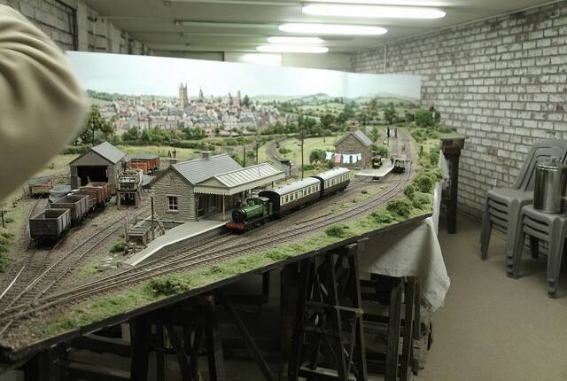 The club's OO gauge layout of Wantage Road station: a green tank engine and two coaches at the platform, the stone station building and canopy, the goods yard and open wagons to the left, and a painted backscene of the town behind.